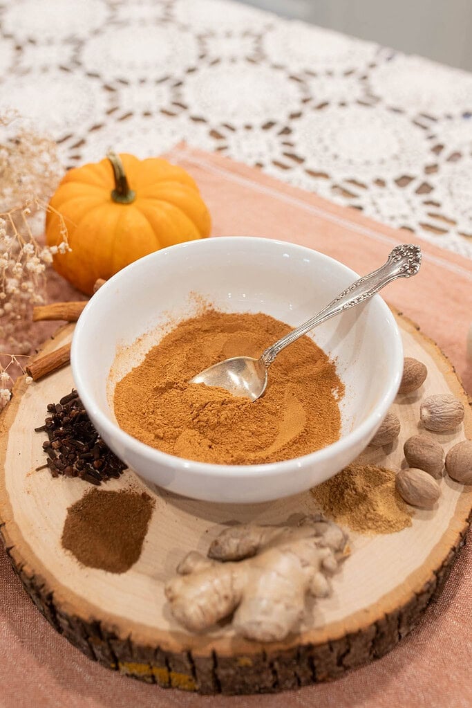 White bowl filled with homemade pumpkin pie spice on a wooden slab surrounded by whole ginger, nutmeg, cloves, cinnamon sticks, and a mini pumpkin on a lace tablecloth