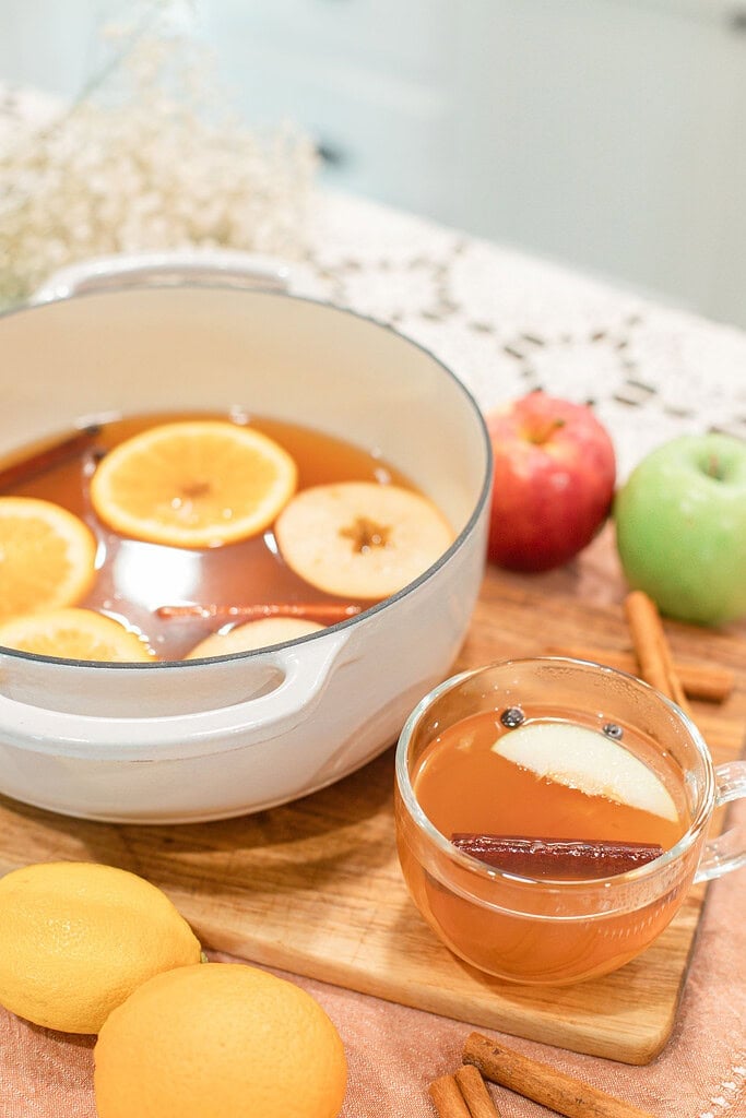 White Dutch oven filled with hot apple cider, orange slices, and a cinnamon stick beside a glass mug of cider garnished with an apple slice, surrounded by lemons, red and green apples, and cinnamon sticks on a wood cutting board