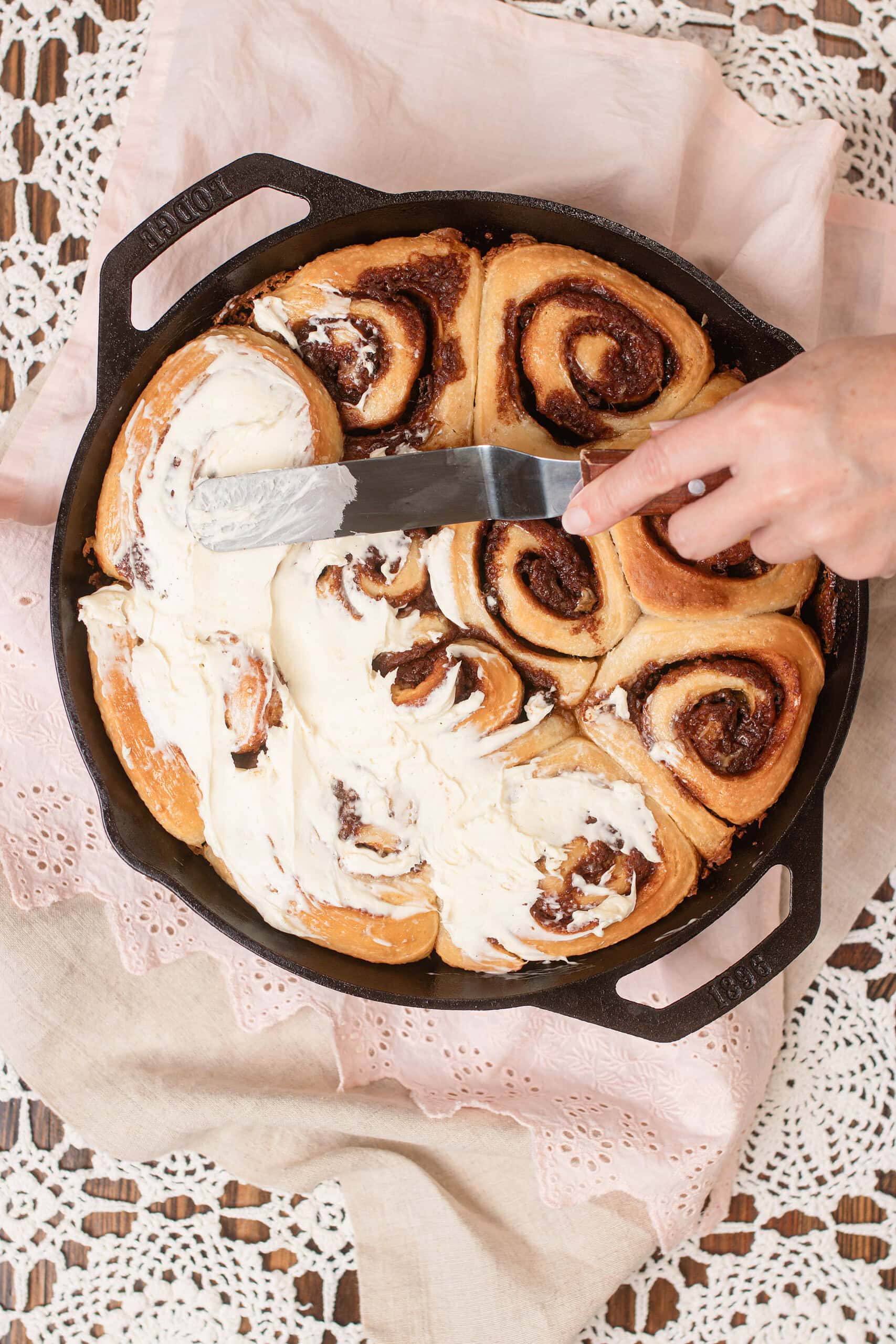 Hand spreading cream cheese frosting over freshly baked sourdough cinnamon rolls in a Lodge cast iron skillet with an offset spatula, on a pink eyelet linen and lace doily