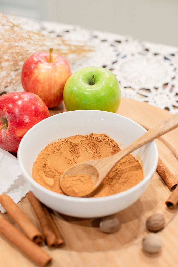 A white bowl of blended homemade apple pie spice with a wooden spoon, surrounded by red and green apples, cinnamon sticks, and whole nutmeg on a wood cutting board with a lace doily.