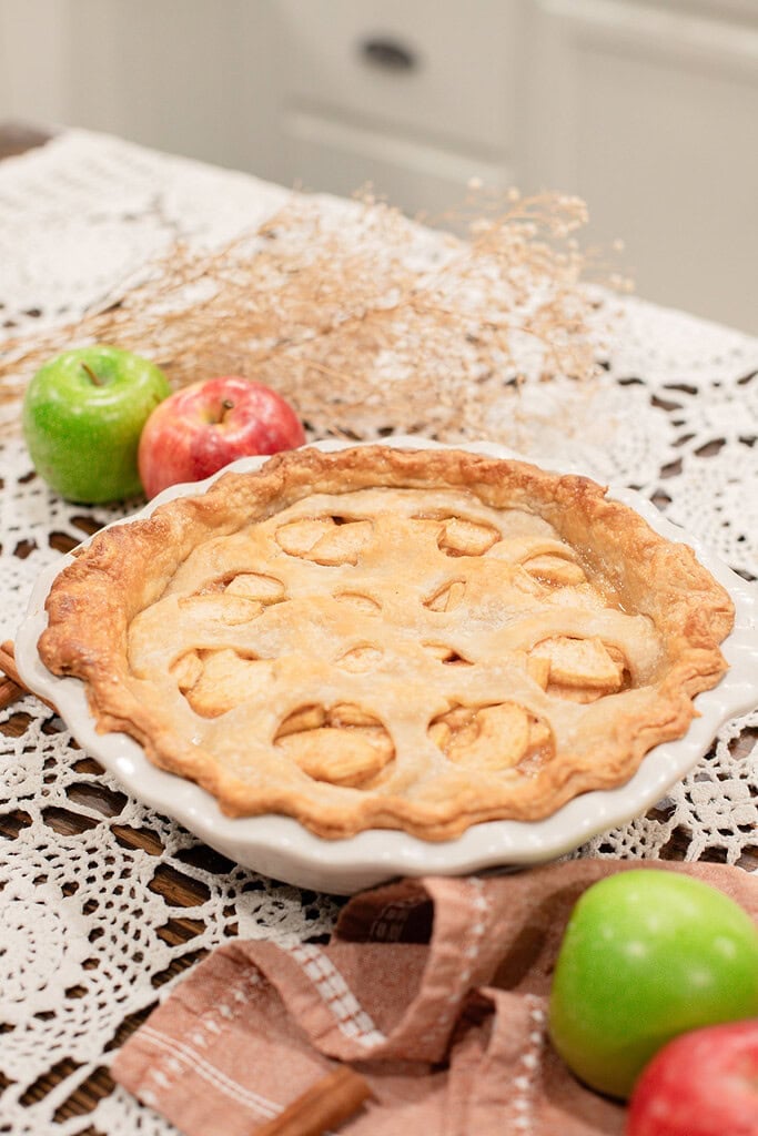 A whole baked sourdough apple pie with apple-shaped cutouts in the top crust in a white ceramic pie dish, styled on a lace doily with red and green apples, dried botanicals, and a dusty rose linen.