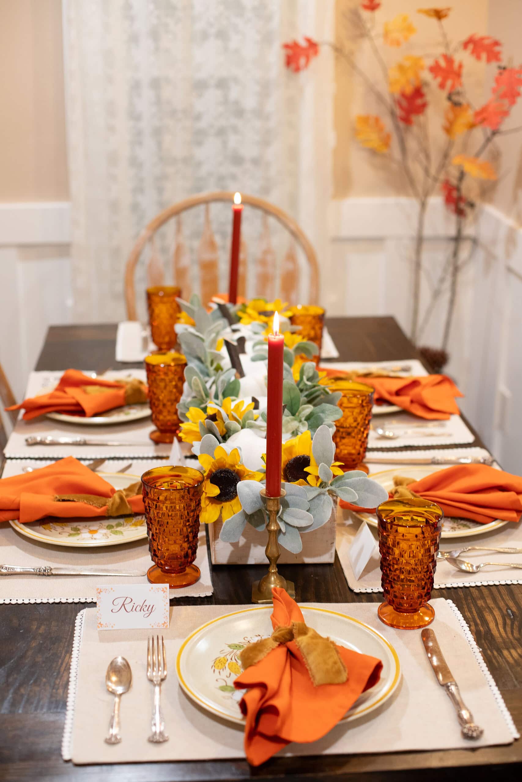 Full-length view of a cozy cottage fall tablescape set for six on a dark wood dining table, featuring orange linen napkins tied with velvet ribbon, amber hobnail glassware, vintage floral dinner plates on cream placemats, a wood box centerpiece filled with white pumpkins, sunflowers, and lamb's ear, lit red taper candles in brass candlesticks, a fall place card, and a colorful autumn leaf branch arrangement in the background.