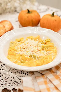 A white bowl of pumpkin mac and cheese with mixed yellow and green pasta in a golden pumpkin cheese sauce, topped with freshly grated parmesan, styled with two small orange pumpkins, a toasted roll, and dried baby's breath on a gold gingham linen.
