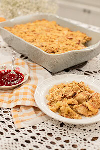 A serving of sourdough cornbread dressing on a white plate alongside a small vintage floral dish of cranberry sauce, with the full baking dish of dressing on a gold gingham linen and lace doily in the background.