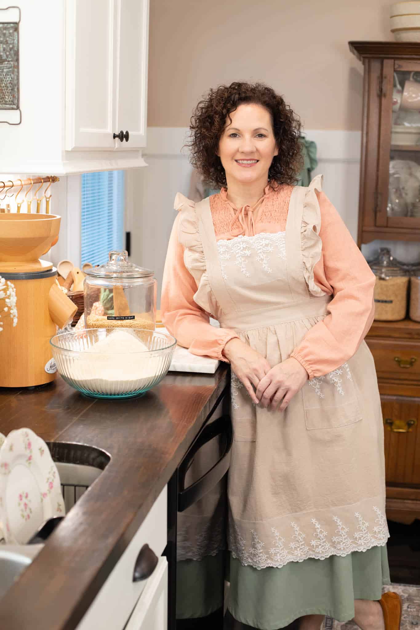 Woman wearing a ruffled apron over a peach blouse leans on a kitchen counter next to a grain mill, wheat berries, and a bowl of fresh flour, smiling warmly.