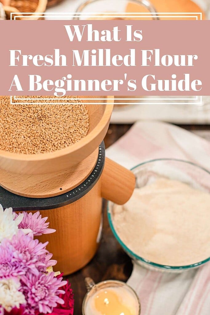 A wooden grain mill loaded with whole wheat berries next to a glass bowl of freshly milled flour, styled with pink and white chrysanthemums and a striped linen, with a title text overlay reading What Is Fresh Milled Flour A Beginner's Guide.