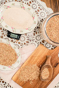Overhead view of whole wheat berries and freshly milled flour on vintage rose-rimmed china plates next to a kitchen scale, with a glass jar of wheat berries and a wooden scoop on a cutting board on a lace doily.